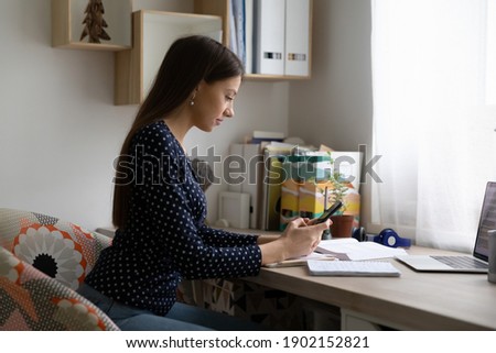 Sudden call. Young lady student distracted from remote study work making answering videocall by phone. Female freelancer spend time at home office read documents check data online via mobile internet