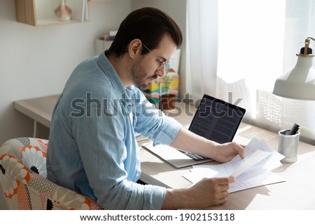 Homesourcing. Concentrated young man sit by laptop pc at home office doing paperwork reading document hardcopy. Remote worker employee analyzing statistic data correcting mistakes in printed report Royalty-Free Stock Photo #1902153118