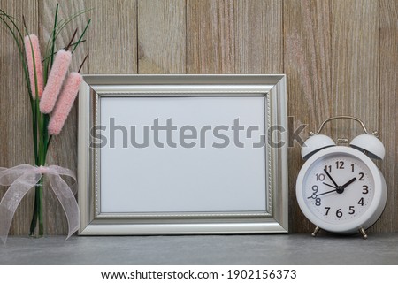 Silver frame mockup with white alarm clock and flower in vase against wood wall for art or copy space.  Spring, springtime, Daylight Saving Time, Easter, birthday, sale, party, event mockup.