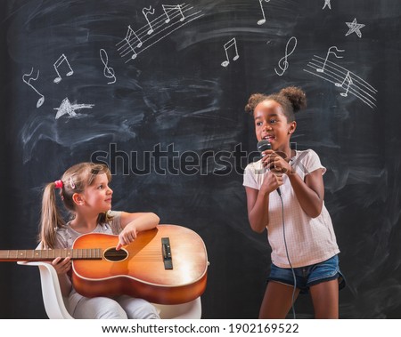 Two beautiful little girls standing in front of a chalkboard in classroom singing and playing the guitar while in music class with musical note symbols drawn on the blackboard