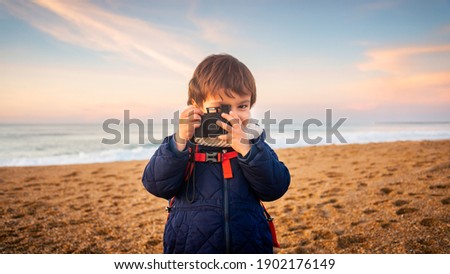 Little boy pretending to take a picture with compact camera on beach at sunset, winter season.
