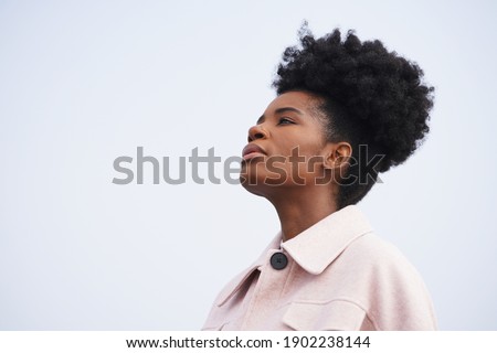Beautiful black woman wearing a pink jacket stands against a stormy blue sky and looks to the left and up in a powerful pose with and updo pony tail afro hairstyle                               Royalty-Free Stock Photo #1902238144