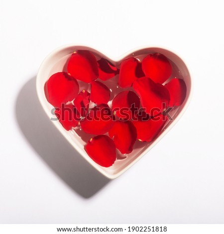 White heart shaped bowl with red rose petals on the water surface. White background