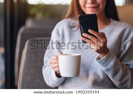 Closeup image of a beautiful young asian woman holding and using mobile phone while drinking coffee