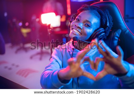 Streamer African beautiful girl shows heart sign with hands professional gamer playing online games computer, neon color.