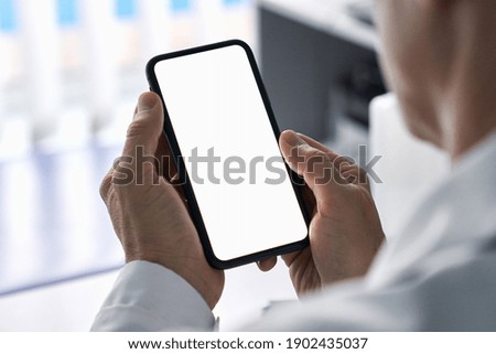 Male doctor holding cell phone in hands using blank white mockup screen technology ehealth mobile app for medical healthcare telemedicine ads, e telehealth online applications. Over shoulder view Royalty-Free Stock Photo #1902435037