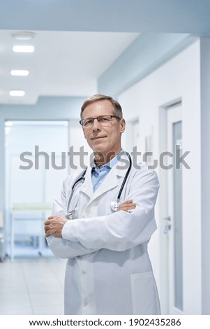 Happy old 60s doctor therapist standing arms crossed in hospital looking at camera. Smiling middle aged male medical professional chief physician, confident mature cardiologist practitioner portrait. Royalty-Free Stock Photo #1902435286