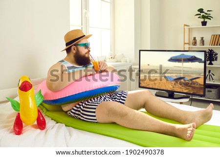 Funny young man with sunglasses and inflatable beach toys sipping cocktail and watching travel show on TV. Concept of canceled summer holiday plans, vacation in lockdown at home or Covid-19 quarantine Royalty-Free Stock Photo #1902490378
