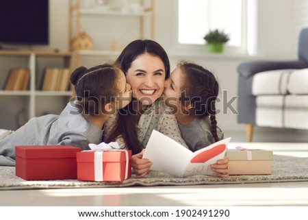 Celebrating Mothers day or Womans day at home concept. Happy young mother woman lying on floor between kissing her daughters and reading handmade postcard for mom on holiday