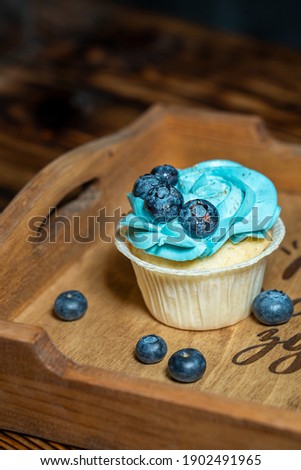 Closeup view of single sweet homemade cupcake or fairy cake decorated with blue creamy cheese topping and blueberries on wooden serving platter. Vertical orientation image, selective focus