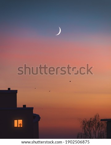 Dramatic sunset colors and young moon at the evening sky. Trees, birds and houses silhouette at night. Light in one lonely window. Cozy nightfall at city under crescent moon. Home family atmosphere. Royalty-Free Stock Photo #1902506875