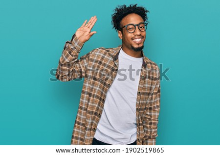 Young african american man with beard wearing casual clothes and glasses waiving saying hello happy and smiling, friendly welcome gesture  Royalty-Free Stock Photo #1902519865