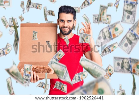 Young handsome man with beard holding delivery cardoboard with italian pizza doing ok sign with fingers, smiling friendly gesturing excellent symbol