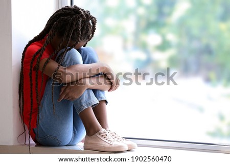 Sad African-American girl sitting on windowsill. Stop racism Royalty-Free Stock Photo #1902560470