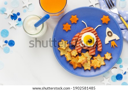Cute smiling alien shaped hamburger served with roast potatoes, carrots and eggs for a healthy lunch or dinner with yoghurt and orange juice. Fun food for kids