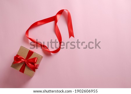 Selective focus of gift box with red ribbon and love shaped paper over pink background; top view flat lay.