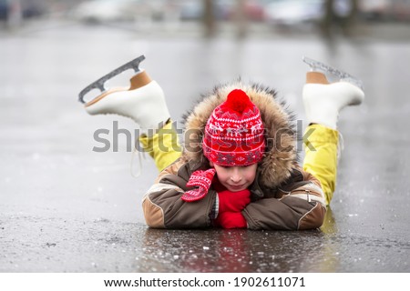 A child in figure skates in winter lies on the ice and smiles.