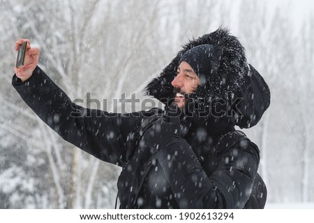 hipster man with a beard taking a selfie with his mobile with a coat to protect himself from the cold while a  snow is falling in a park with snowy trees in the background in winter. selective focus
