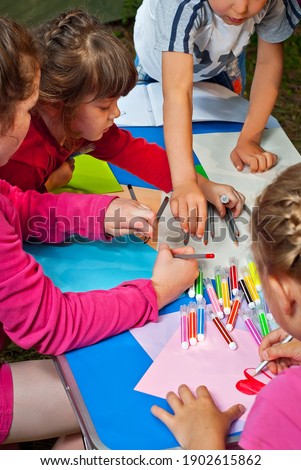 Children draw in nature. Lots of colored papers and pencils on a blue table. Boys and girls play outdoors. Drawing lesson in the park. Rest in a tent camp in the summer.