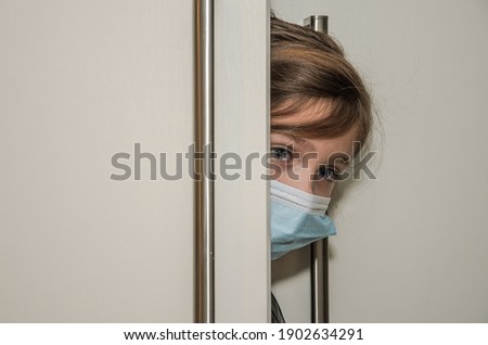 Little girl child in medical mask peeks out of the door	
