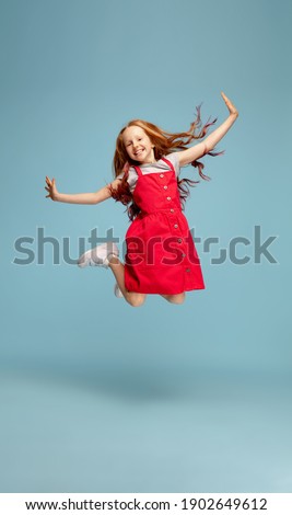 Weightless, freedom. Happy redhair girl in red dress on blue studio background. Looks happy, cheerful. Copyspace for ad. Childhood, education, emotions, facial expression concept. Jumping high, flying