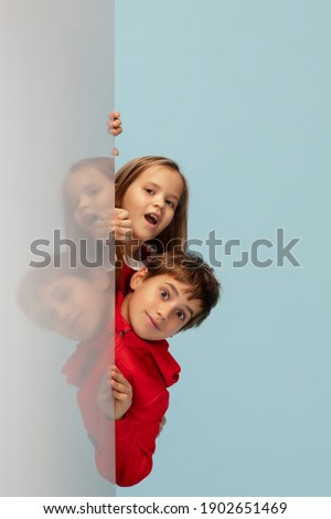 Looking up. Happy children isolated on blue studio background. Look happy, cheerful. Copyspace for ad. Childhood, education, emotions, facial expression concept. Peeking out from behind the wall.