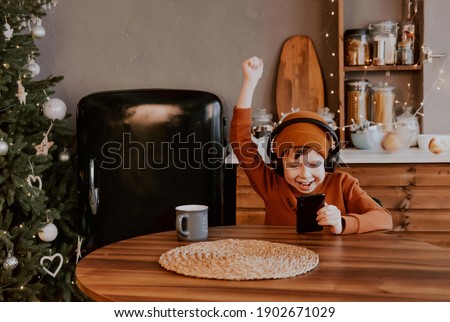 A boy in the kitchen listens to music through wireless headphones. Watch the movie over the phone. Games in the phone. Winning an online game. Vacation at home. Fashionable boy listens to music. 