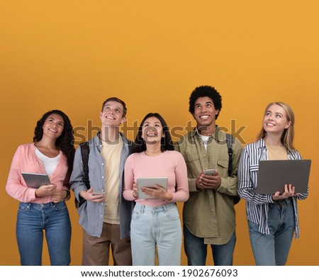 Great offer for students, important information and ad. Smiling digital young international people with backpacks, gadgets looking up, isolated on orange background, studio shot, empty space, square