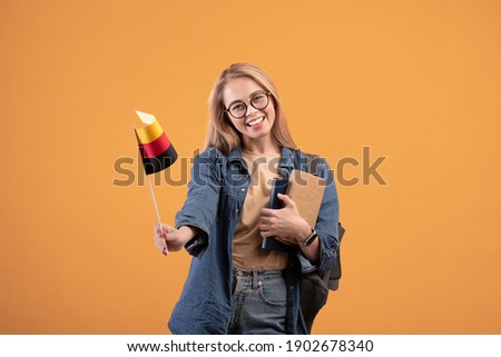 Tourist traveling, trip and learning Deutsche language. Teen happy female in glasses with notebooks and backpack waving small flag of Germany, isolated on beige background, studio shot, empty space Royalty-Free Stock Photo #1902678340