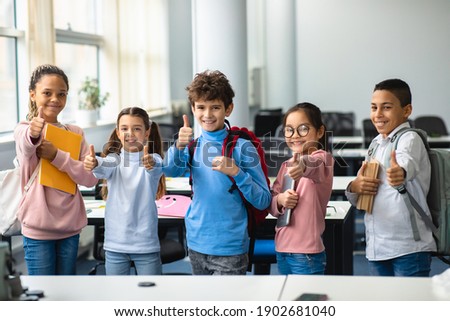 Education Concept. Diverse group of happy smiling international classmates standing in line with notebooks and backpacks in classroom and showing thumbs up sign gesture, enjoying studying at school