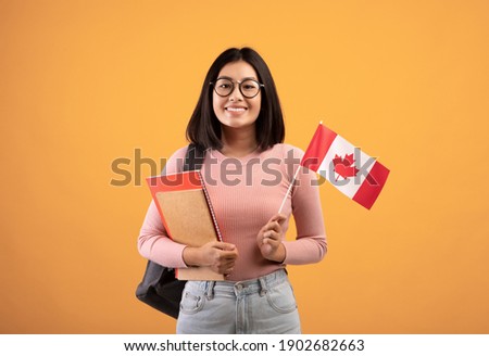 Travel, modern education and student exchange. Young cheerful asian woman in glasses with notebooks, backpack holding small flag of Canada, isolated on beige background, studio shot, empty space Royalty-Free Stock Photo #1902682663