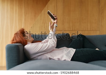 Young happy woman lying on the sofa after hard work day and making a video call using smartphone and earphones, smiling and gesturing. Communication with family and friends during quarantine