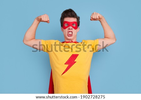 Mad young man in superhero costume looking at camera and demonstrating muscles against blue background