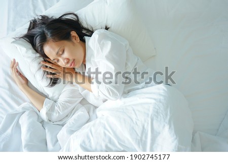 Young Asian woman sleeping in bed and lying on soft pillow, white bedding linen mattress in the morning, smiling happily in shirt, comfort rest at high angle or top view. Royalty-Free Stock Photo #1902745177