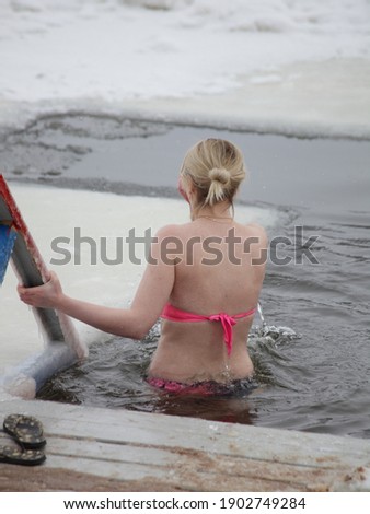 Winter swimming sport, a mature Caucasian woman in a swimsuit enters the ice hole water on a Sunny frosty winter day, healthy lifestyle