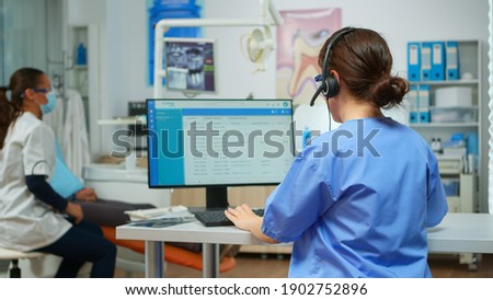 Dentist assistant making appointments using headset sitting in front on computer while doctor is working with patient in background examining teeth problem. Nurse taking notes in stomatological office Royalty-Free Stock Photo #1902752896