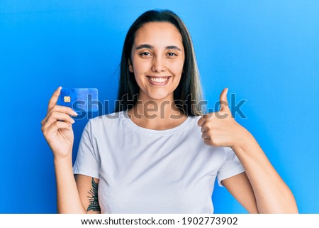 Young hispanic woman holding credit card smiling happy and positive, thumb up doing excellent and approval sign 