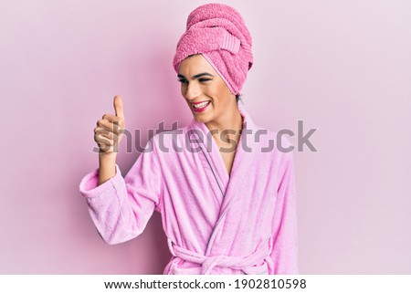 Young man wearing woman make up wearing shower towel on head and bathrobe looking proud, smiling doing thumbs up gesture to the side 