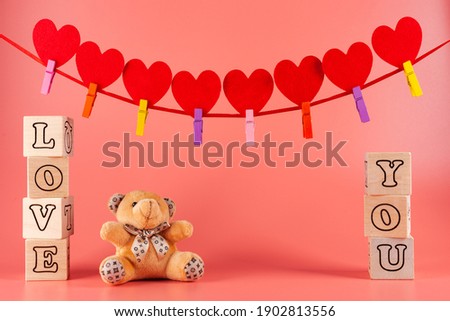 on the coral background at the edges of the frame of wooden cubes with letters laid out the phrase love you, between the cubes sits a small teddy bear, at the top of the frame garland with red hearts