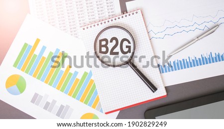 Magnifying glass on B2G text on notepad, dice, spectacles, pen, laptop calculator on wooden table - business, banking, finance and investment concept