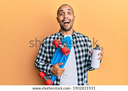 Hispanic adult skater man holding skate and graffiti spray celebrating crazy and amazed for success with open eyes screaming excited. 