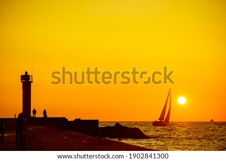 a white sailboat floats against the backdrop of the setting sun and a lighthose away from it near which people are standing