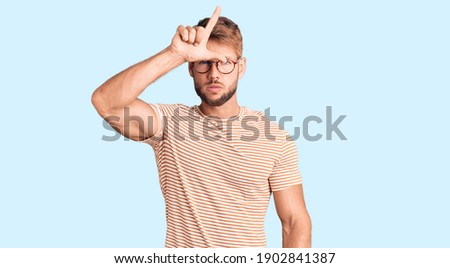 Young caucasian man wearing casual clothes and glasses making fun of people with fingers on forehead doing loser gesture mocking and insulting. 