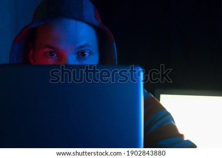 hacker, programmer or gamer in hoodie using laptop in dark room in red blue neon light, hacker gaze into the monitor, cyber security and hacking attack concept