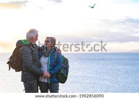 Smiling senior couple with backpack on their back enjoys the hike on the ocean cliffs. On the horizon the profile of an island and a ship.