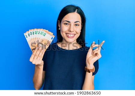 Beautiful young woman holding bunch of 50 euro banknotes cheerful with a smile on face pointing with hand and finger up to the side with happy and natural expression 