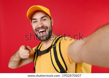 Close up delivery man 20 in yellow cap t-shirt uniform thermal bag backpack doing selfie isolated on red background studio. Fun male employee work as courier Service coronavirus covid-19 virus concept