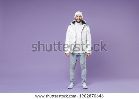 Full length of smiling attractive man in warm white padded windbreaker jacket hat standing looking camera isolated on purple background studio portrait. People lifestyle cold winter season concept