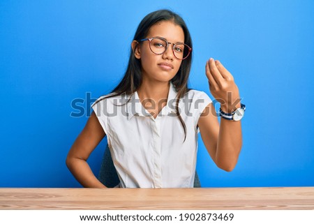 Beautiful hispanic woman wearing casual clothes sitting on the table doing italian gesture with hand and fingers confident expression 
