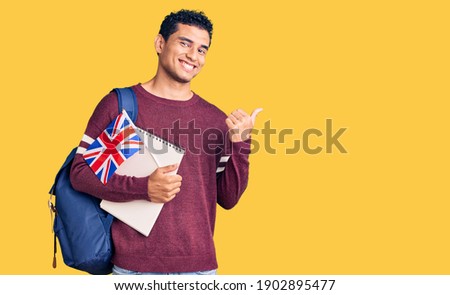 Hispanic handsome young man exchange student holding uk flag pointing thumb up to the side smiling happy with open mouth  Royalty-Free Stock Photo #1902895477